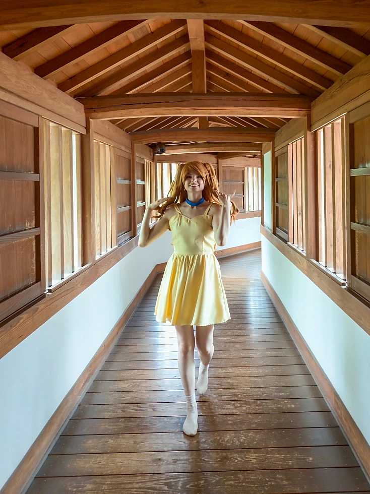 Full-body Asuka Langley cosplay walking through a long wooden castle corridor in a yellow summer dress